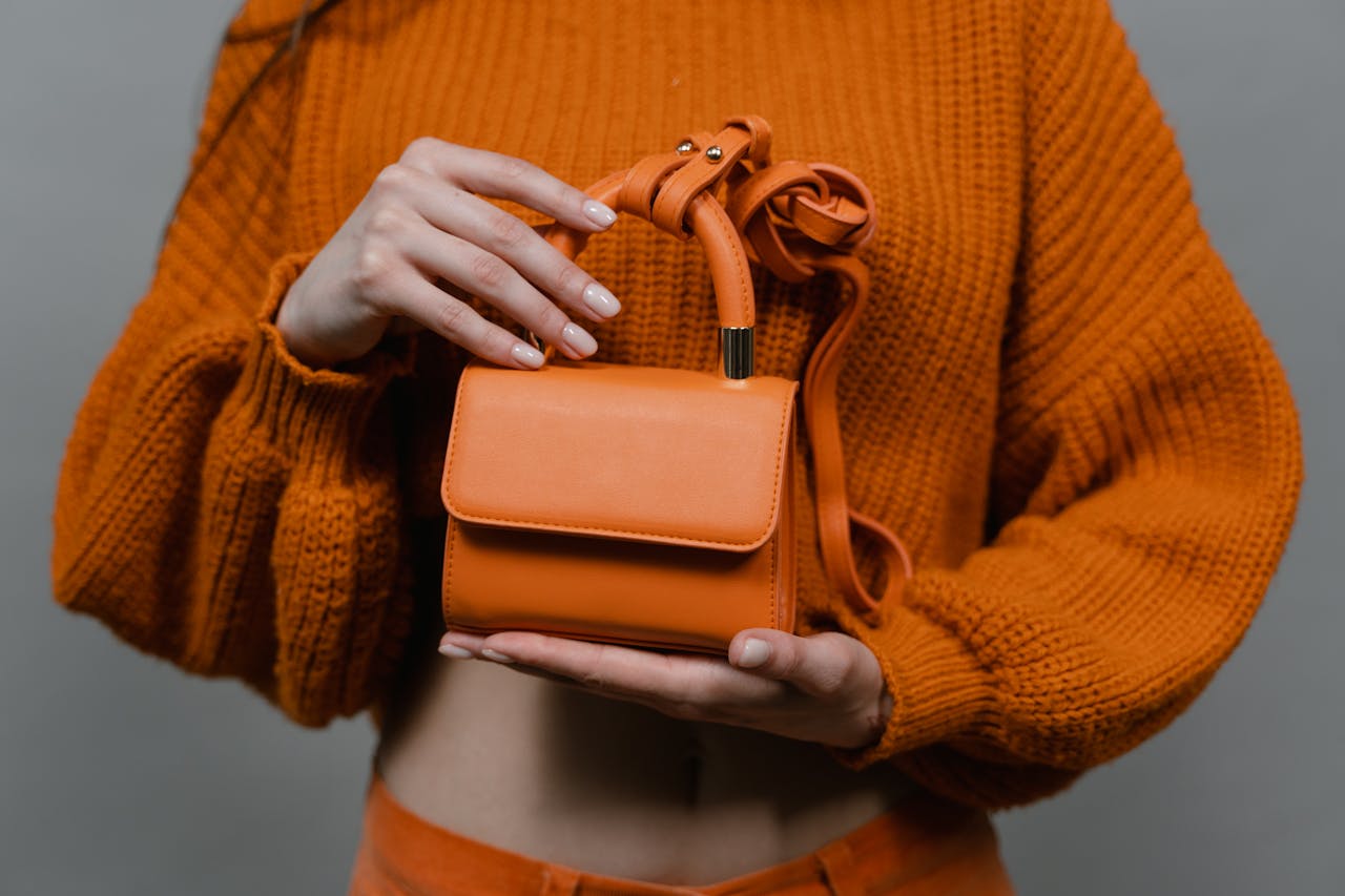 services-03 Woman holding an orange leather mini handbag against an orange sweater in a studio setting.